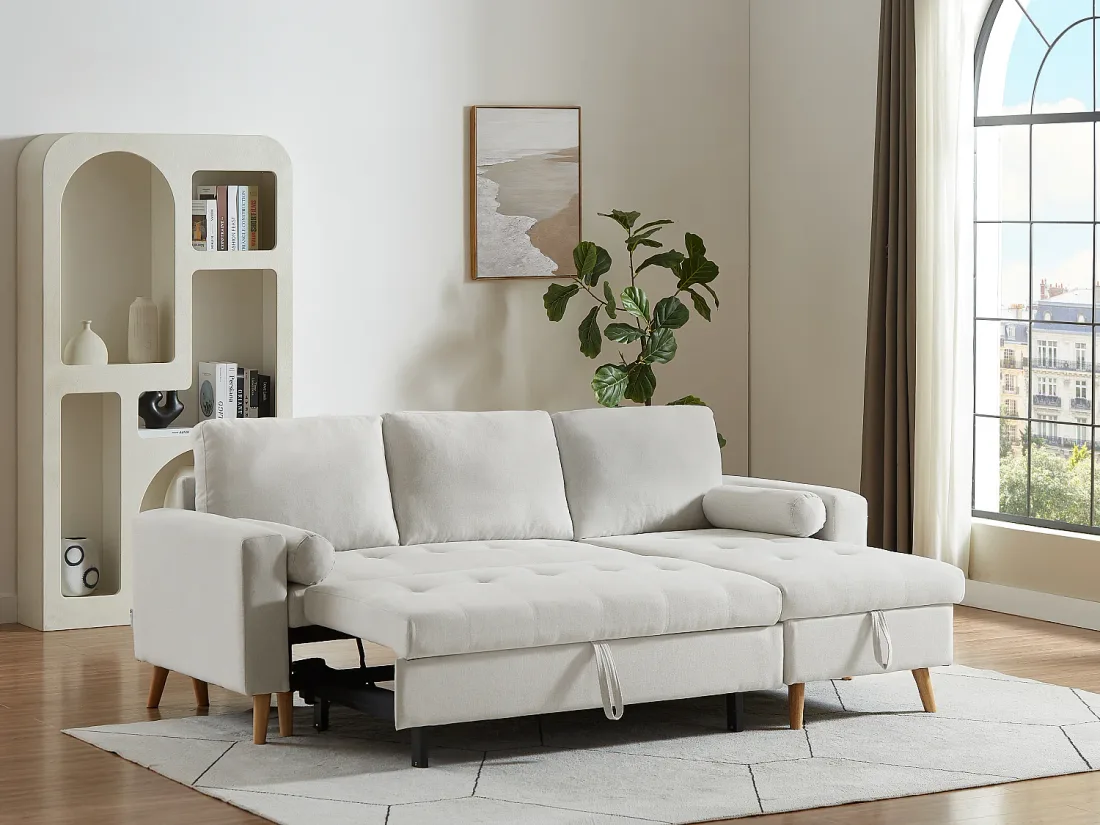 Canapé Kori beige ouvert en lit, matelas capitonné déplié, à côté d'une étagère blanche et d'un ficus.