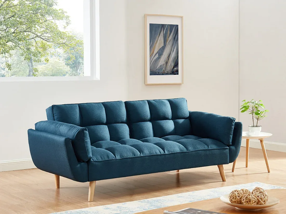 Canapé Loeline bleu en tissu matelassé dans un salon lumineux, près d'une fenêtre, d'un tableau et d'une petite table.